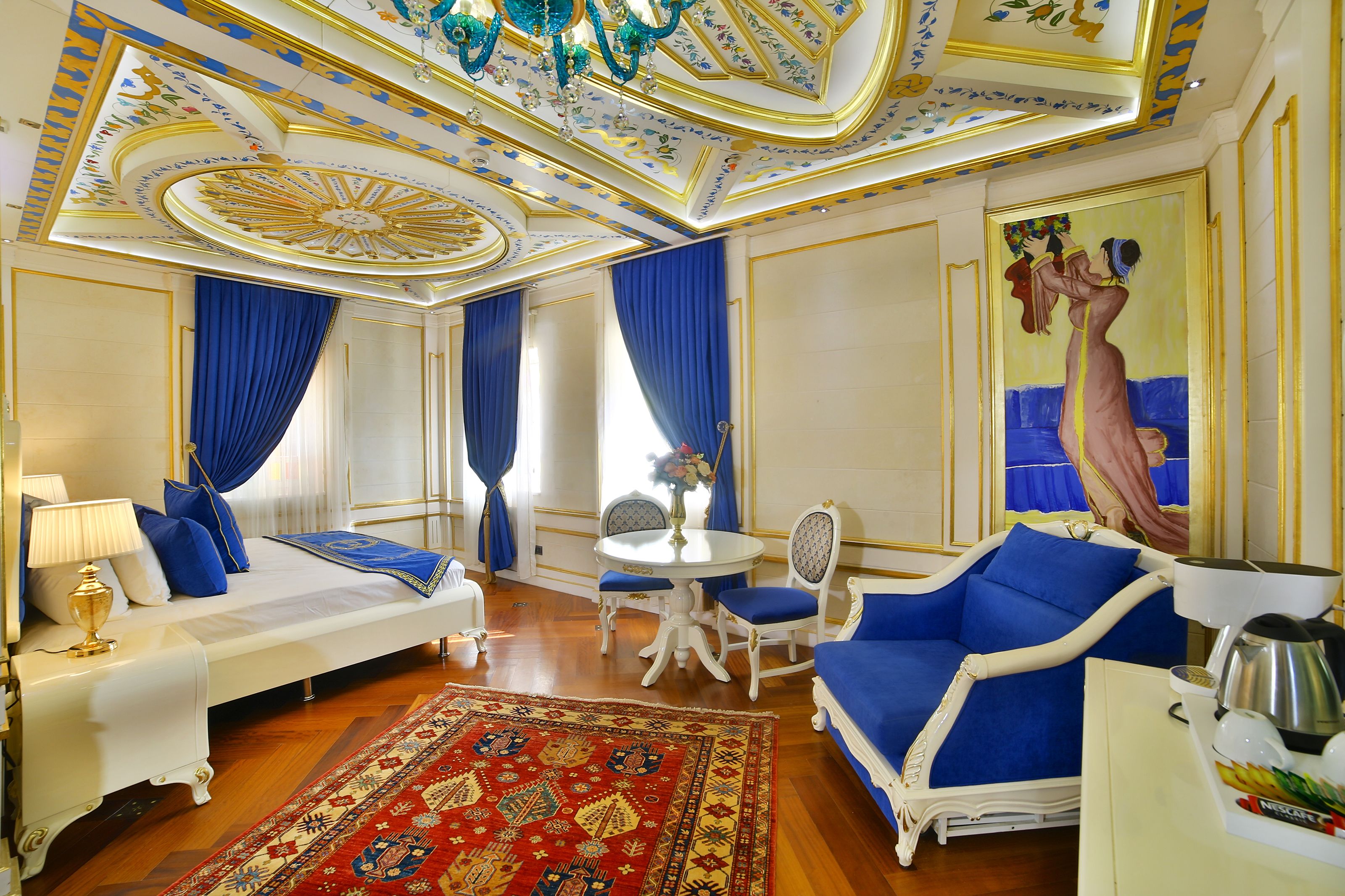 Une chambre luxueuse avec un grand lit, un canapé bleu recouvert de tissu, une petite table ronde avec des chaises et un plafond décoratif.