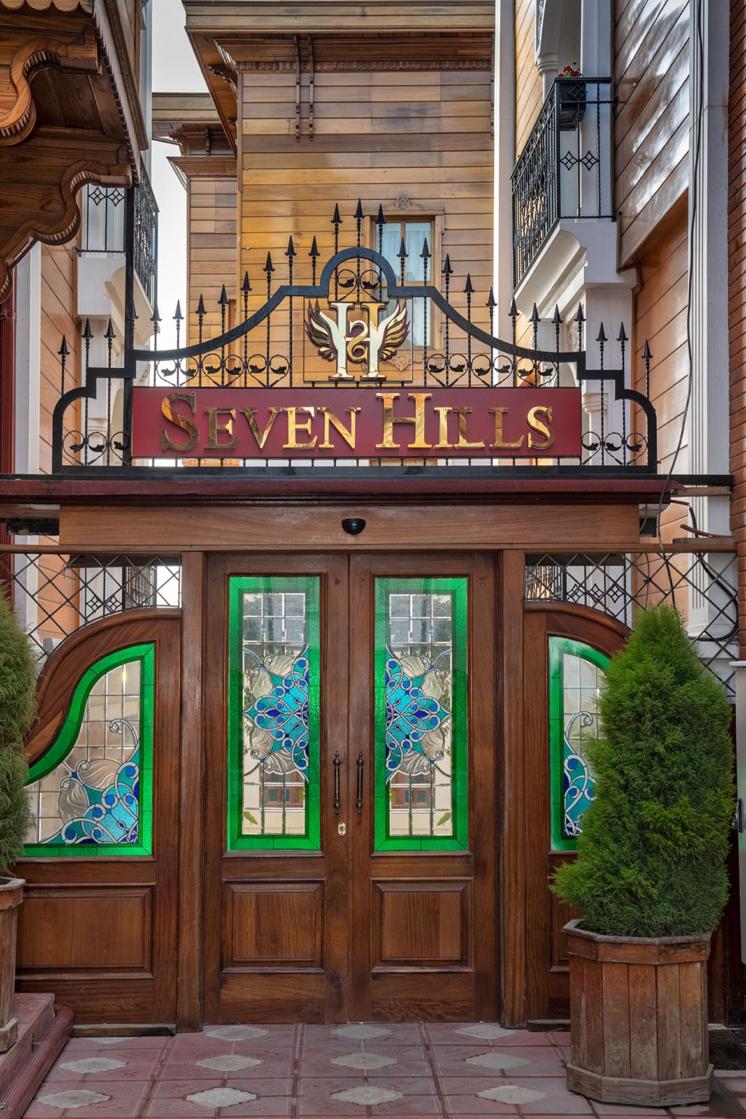 Entrada de hotel con un letrero que dice "SEVEN HILLS", puertas dobles de madera con paneles de vidrio verde y una reja de hierro forjado encima.