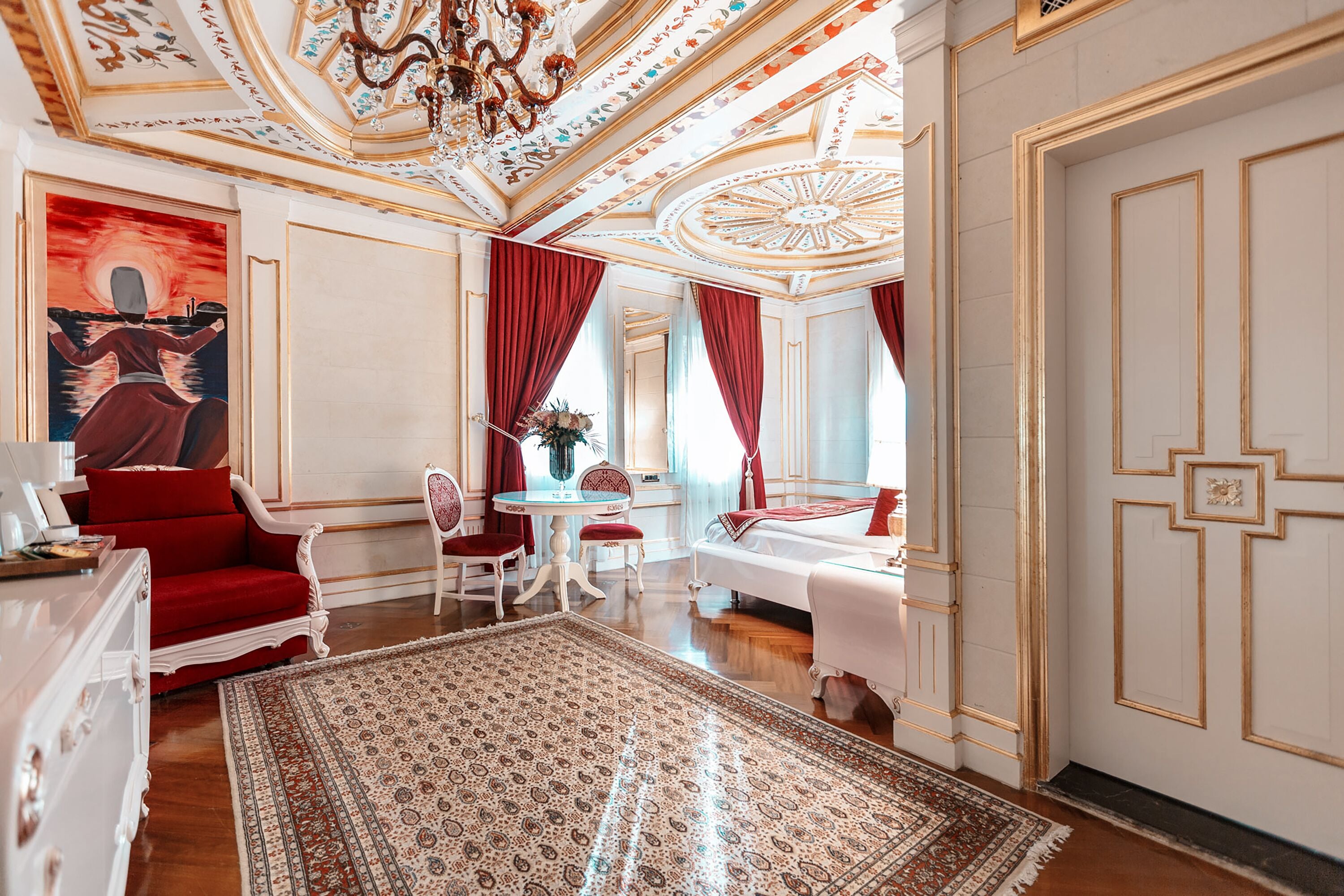 A luxurious room with a large ornate ceiling, a red sofa, a white bed, and a dining area with a table and chairs.