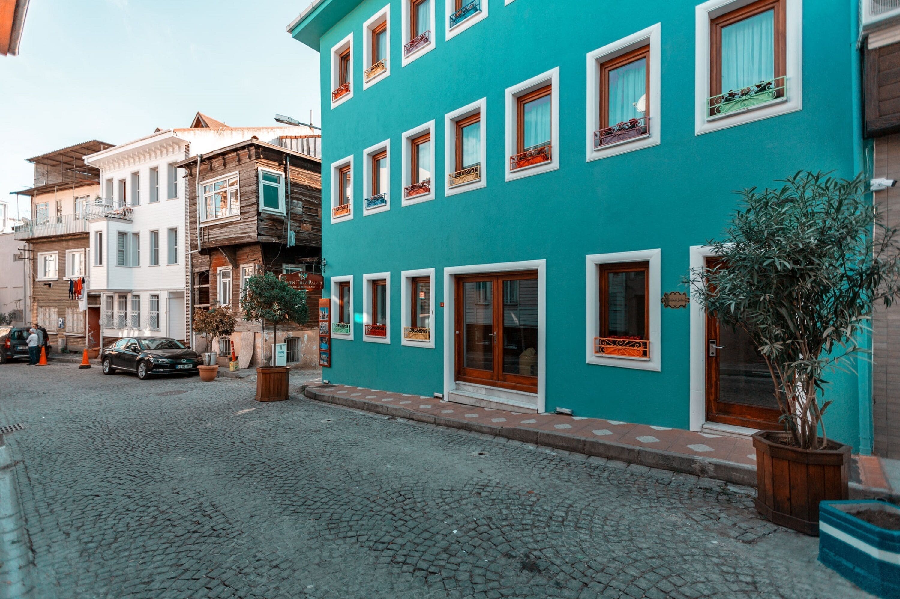 Eine Straße mit Kopfsteinpflaster, ein türkisfarbenes Gebäude mit zahlreichen Fenstern und Balkonen sowie ein geparktes Auto.