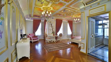 Una habitación lujosa con elaborados adornos en el techo, una araña y muebles elegantes, incluyendo un sofá, sillas y una pequeña mesa.