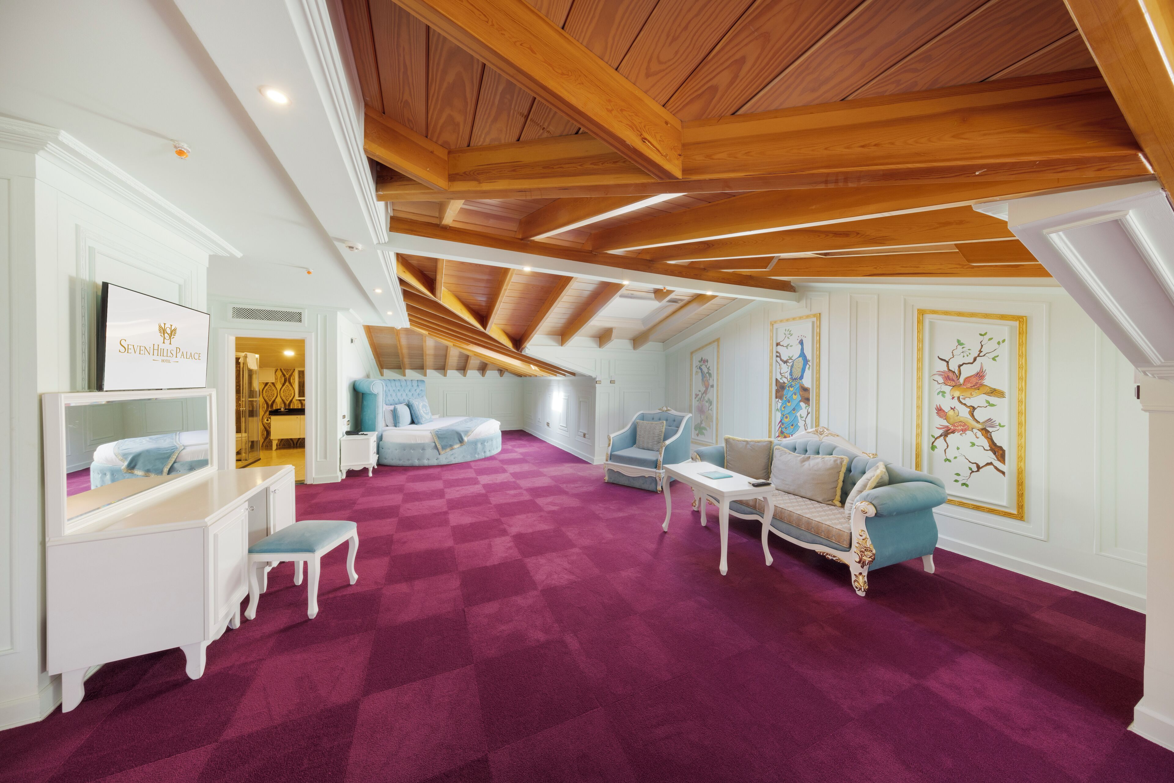 Een ruime kamer met een hoge plafond, houten balken, een geruit tapijt en een zithoek met een bank en fauteuils.