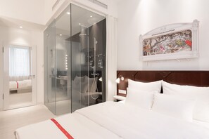 A modern hotel room with a large bed, a glass-enclosed walk-in closet, a shower with a glass door, and a wall-mounted artwork.
