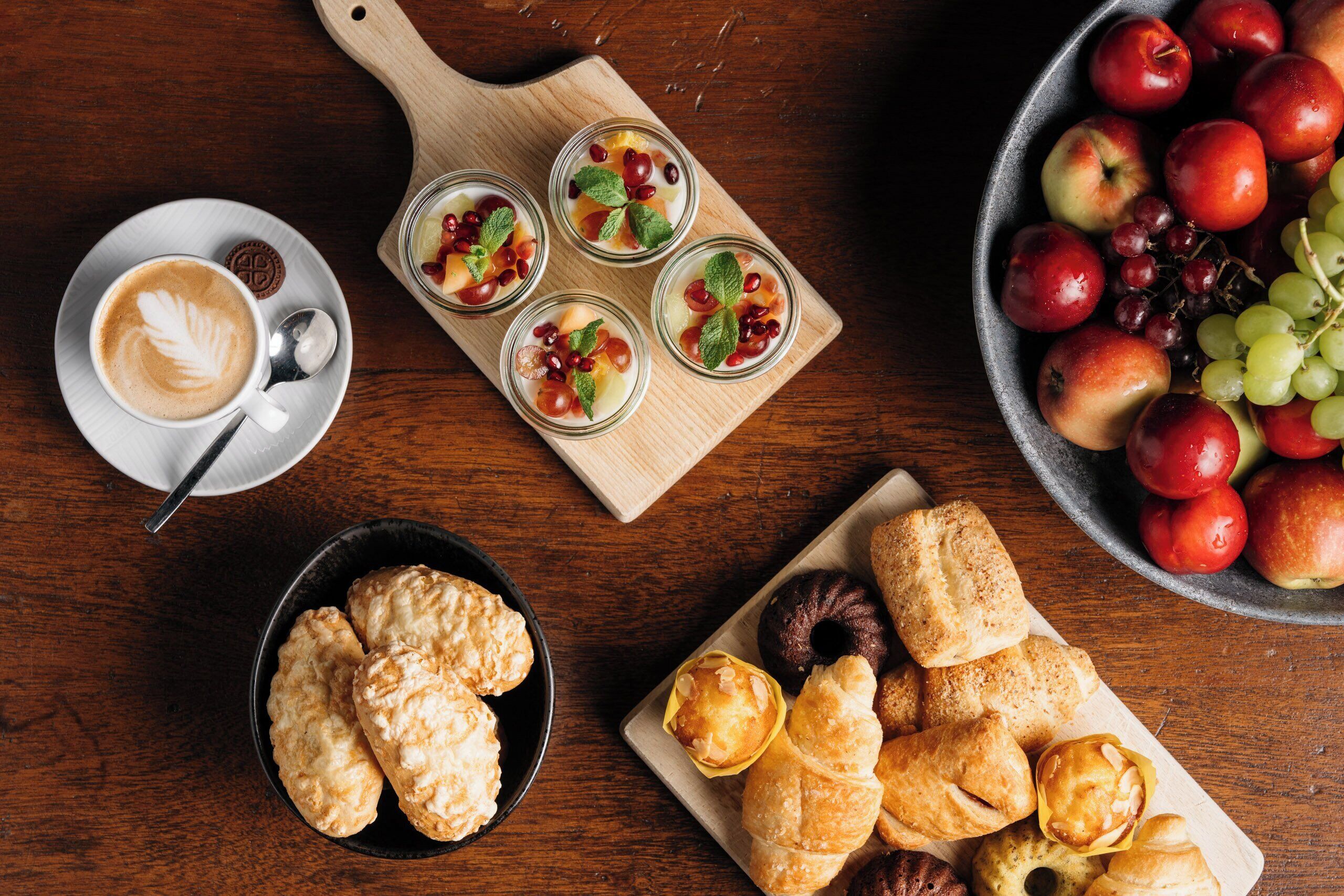 A wooden table with a bowl of apples, a plate of pastries, a cup of coffee, and three glasses of fruit salad.