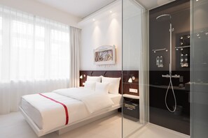 A modern hotel room with a large bed, a glass shower enclosure, and a mounted artwork above the bed.