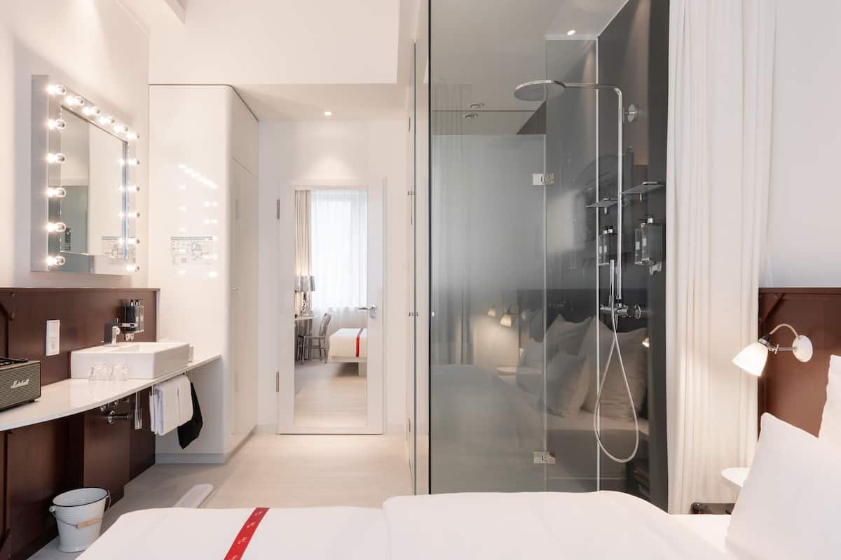A modern hotel room with a large bed, a glass-enclosed shower, a vanity with a mirror, and a hallway leading to a dining area.