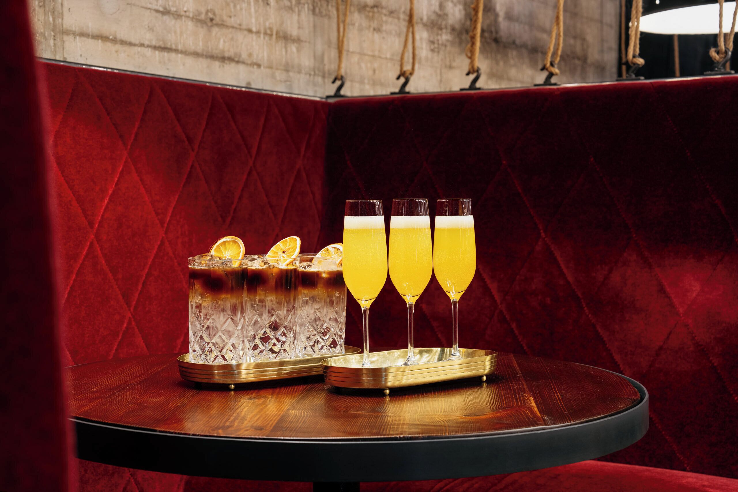 A bar area with a red velvet backdrop, a wooden table, and a glass pitcher with three glasses of a yellow beverage.