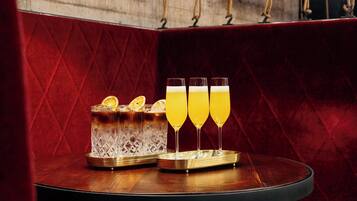 A bar area with a red velvet backdrop, a wooden table, and a glass pitcher with three glasses of a yellow beverage.