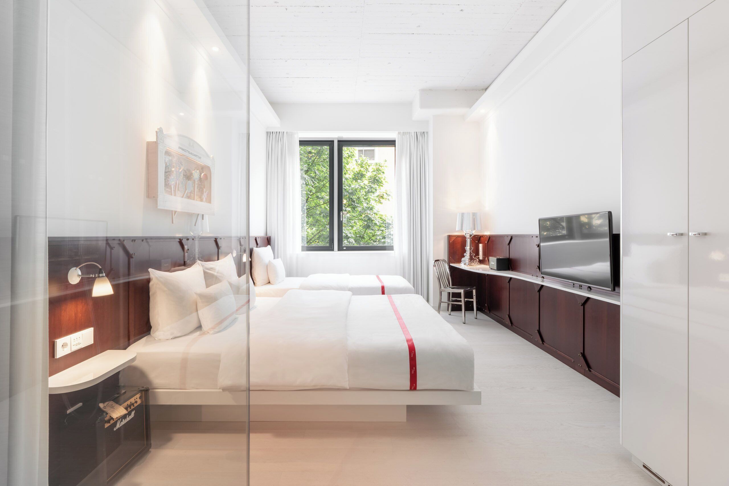 A modern hotel room with a large bed, a desk, a TV, and a window with a view of greenery.