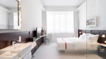 Una habitación de hotel moderna con una cama grande, un tocador con espejo e iluminación, un televisor y un ventanal con cortinas.
