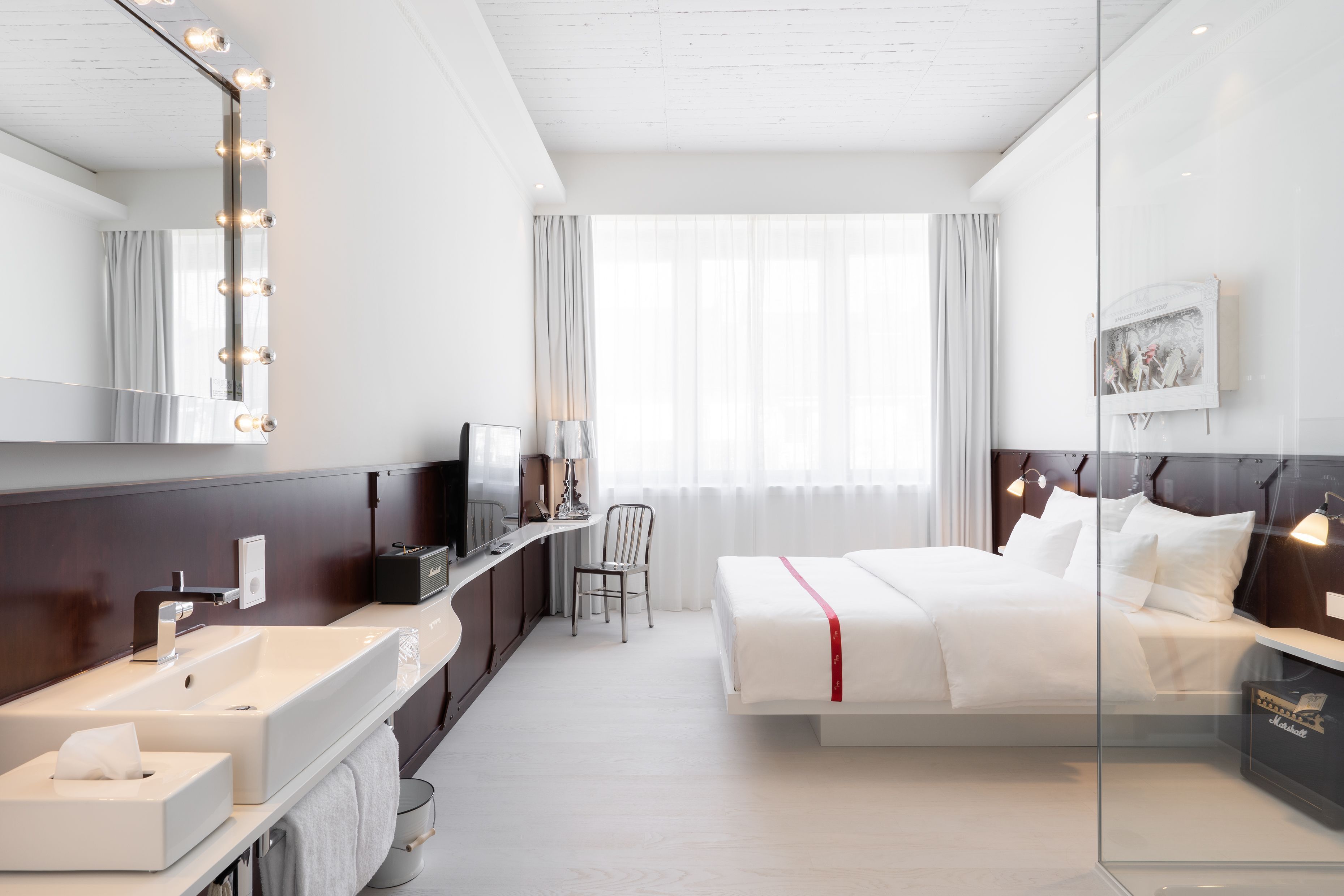 Una habitación de hotel moderna con una cama grande, una tocador con espejo e iluminación, un escritorio con silla y un ventanal con cortinas.