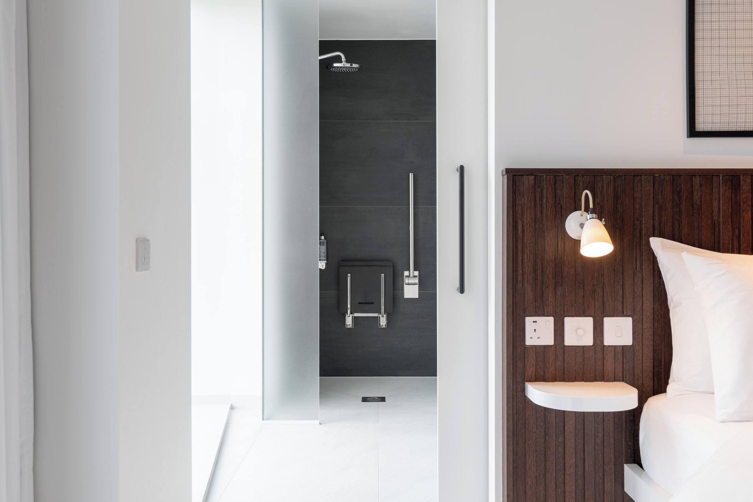 A modern bathroom with a shower, toilet, and a bedside table with a lamp.