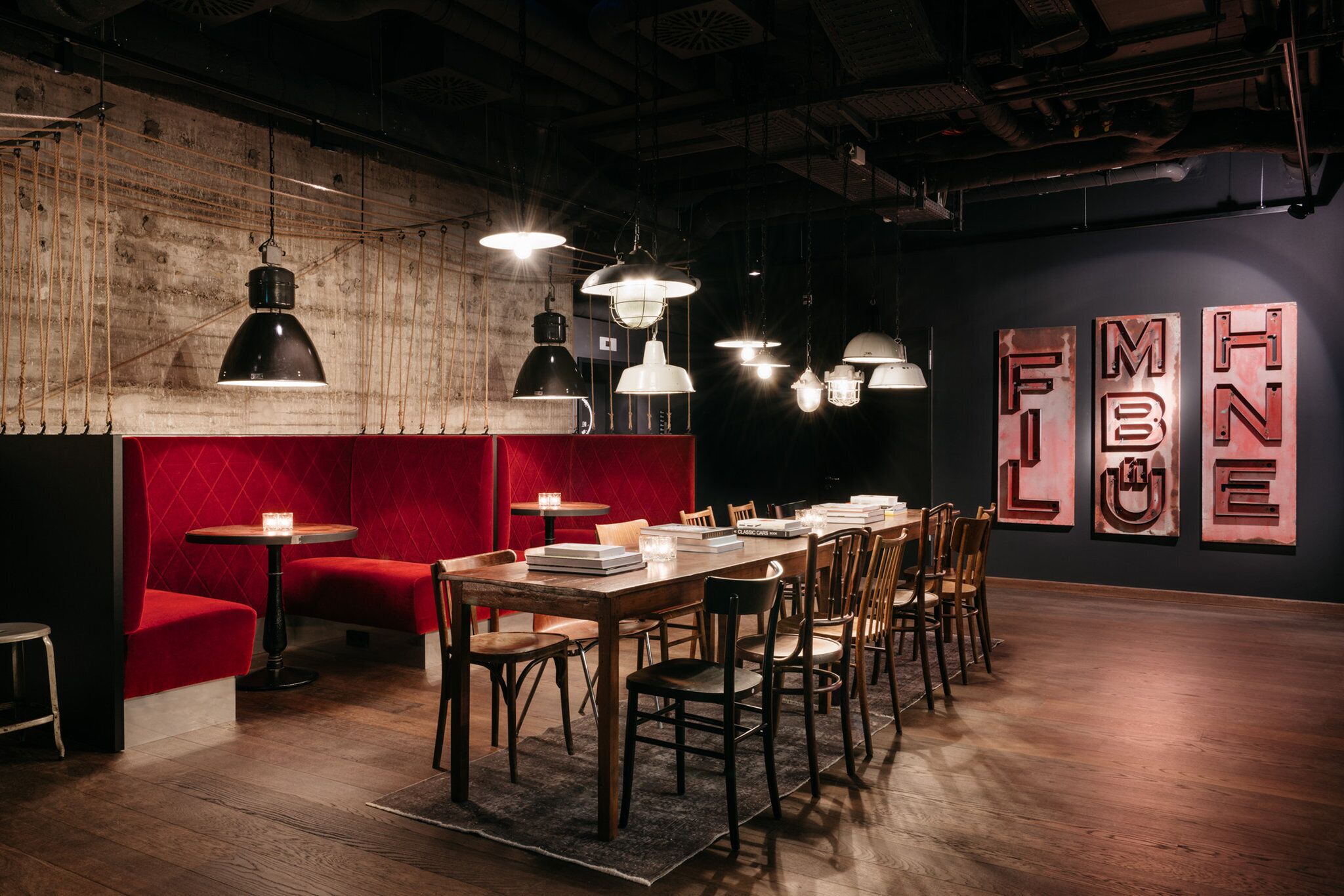 L’intérieur d’un restaurant présente une longue table en bois, des sièges rembourrés de couleur rouge, des suspensions lumineuses et un mur orné de grandes lettres métalliques.