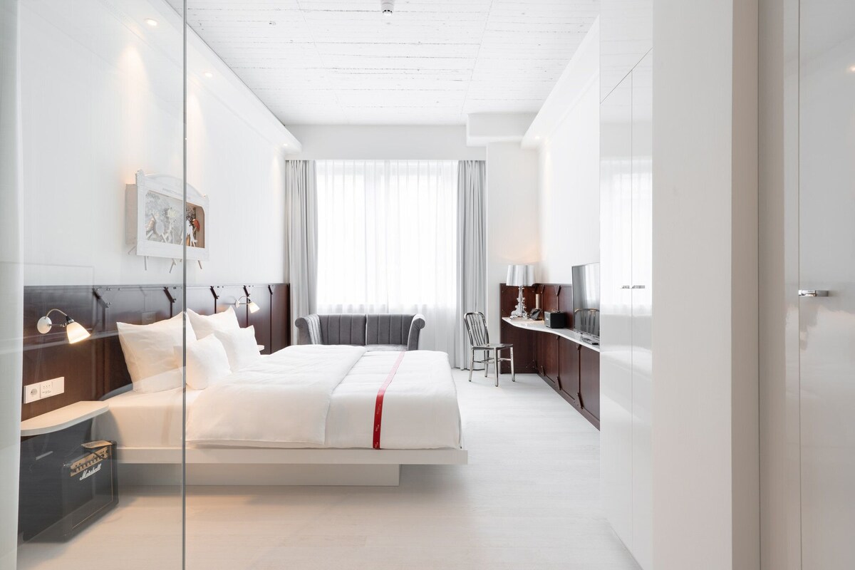 A modern hotel room with a bed, bedside table, lamp, and a view of the city.