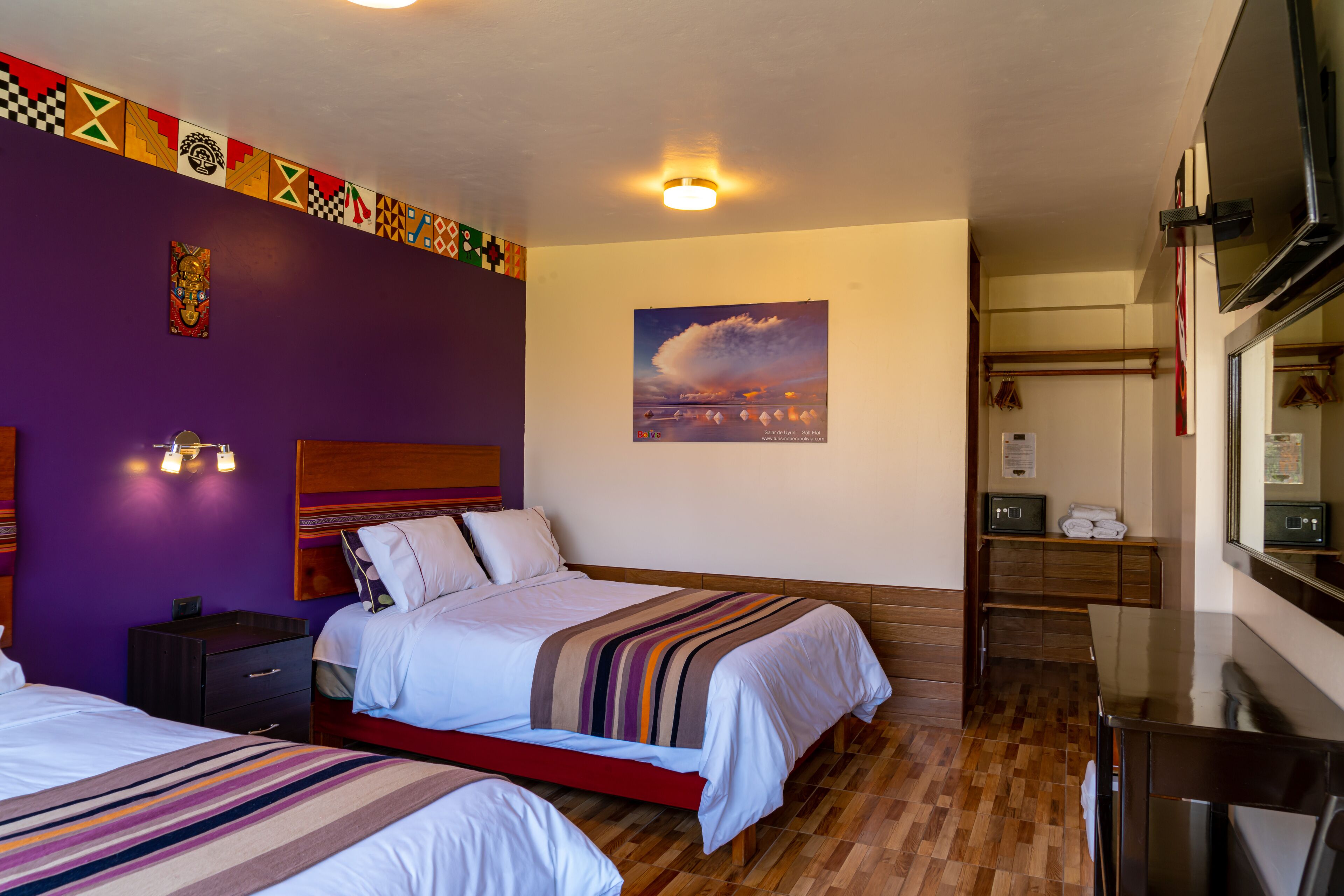A hotel room with two beds, a wooden floor, a purple accent wall, a framed picture, and a small desk with a mirror.