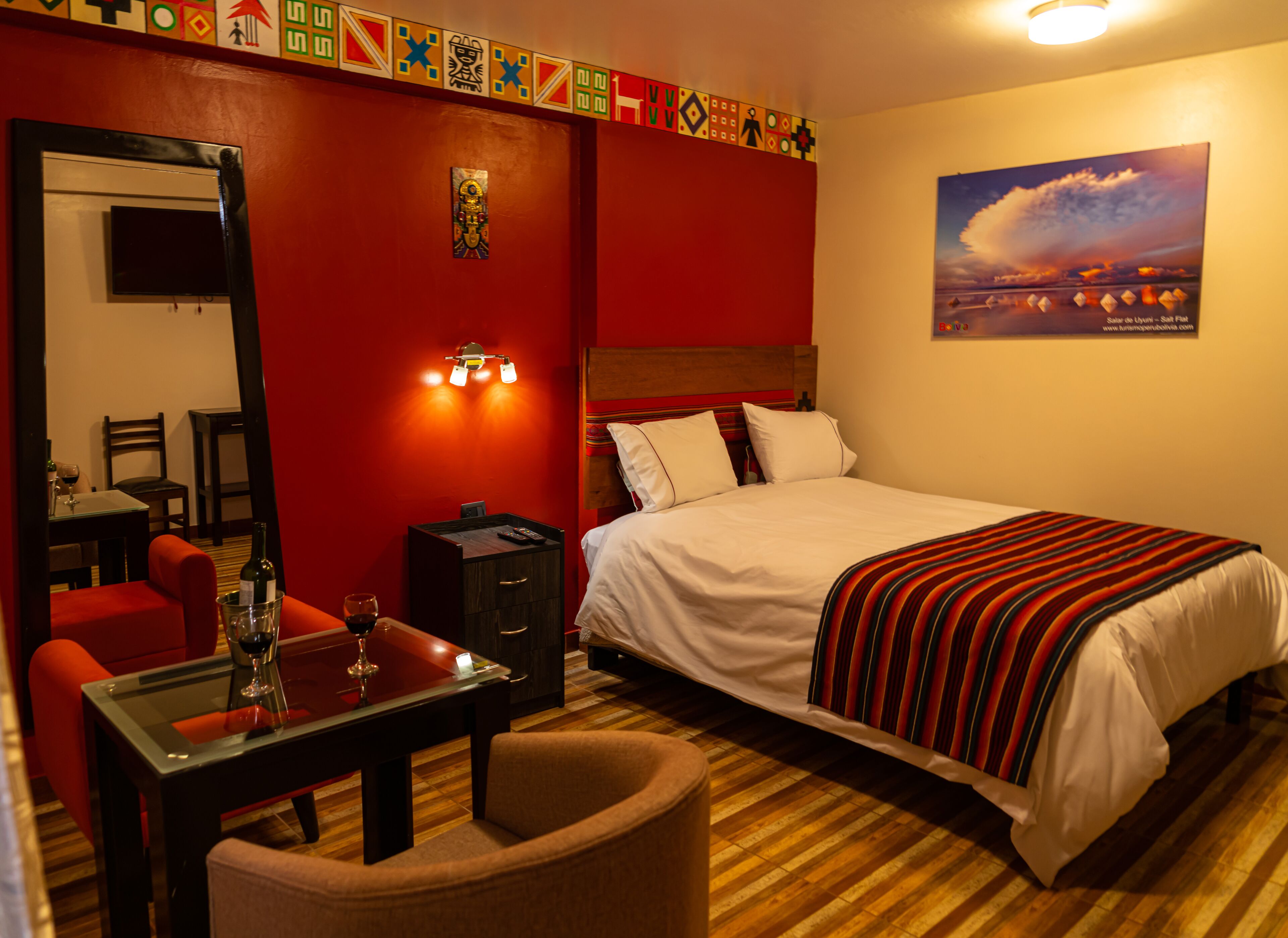 Habitación de hotel con cama, escritorio con silla, pequeña mesa con botella y vaso, mesita de noche y un cuadro en la pared.