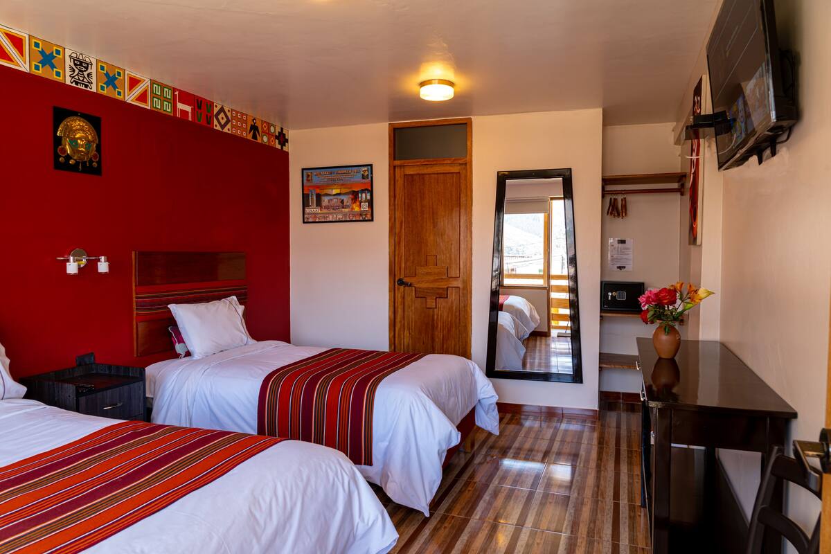A hotel room with two beds, a wooden door, a mirror, a desk, and a vase with flowers.