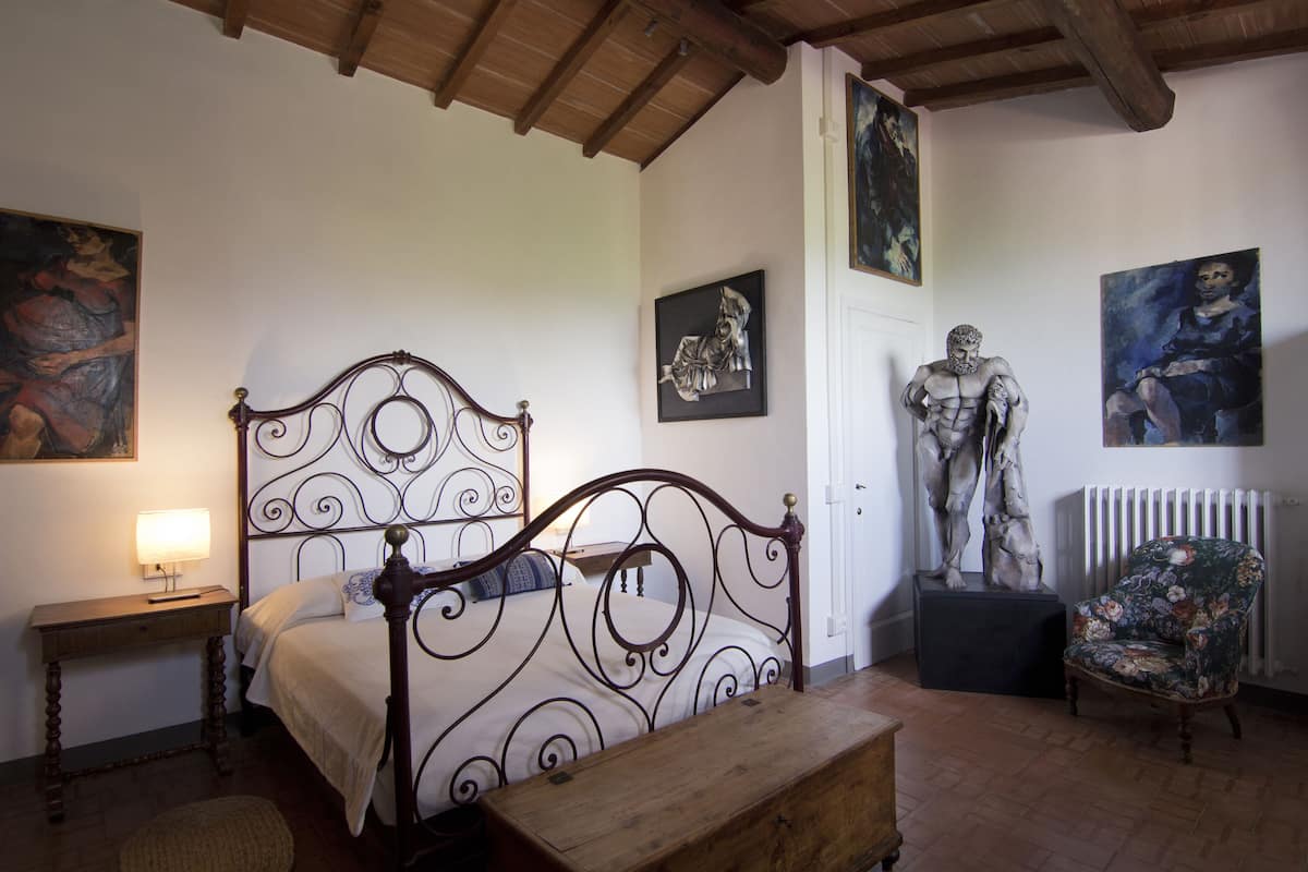 Une chambre à coucher avec un lit en bois, une table de chevet avec une lampe, un banc et une chaise. Les murs sont ornés de tableaux et d’une statue.