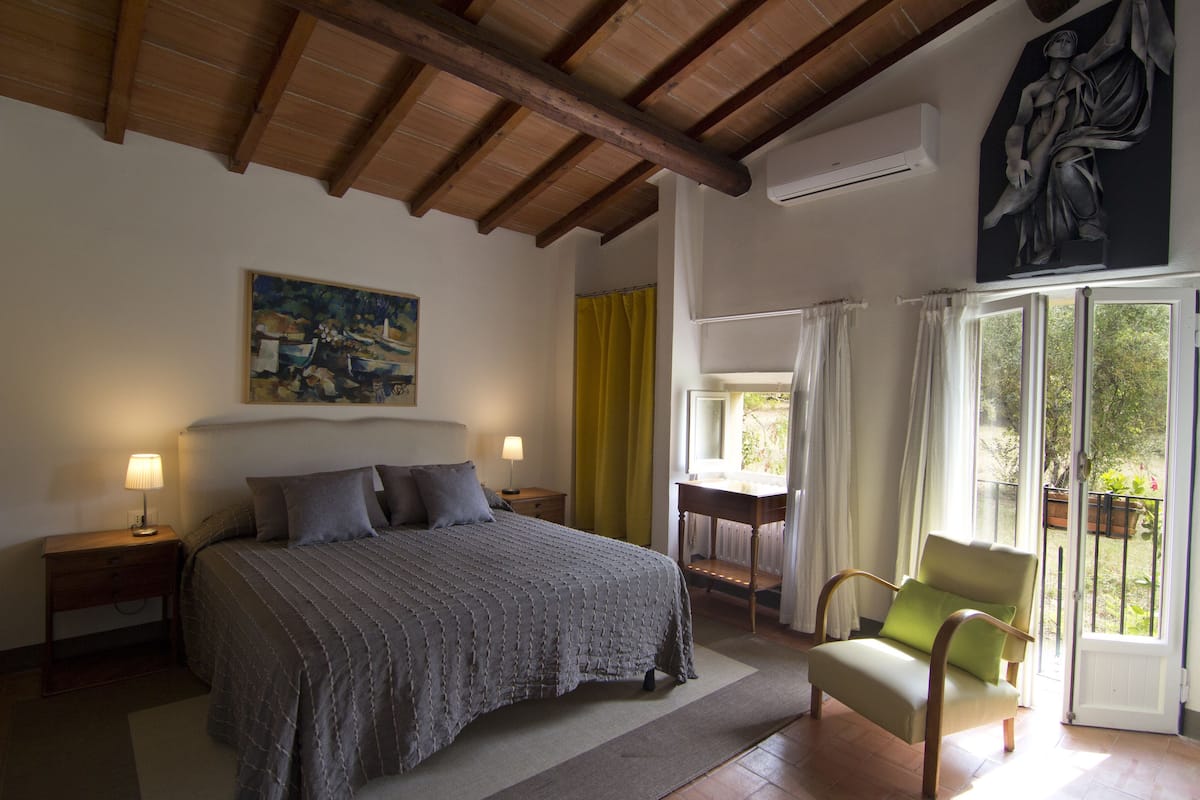 Une chambre à coucher avec un lit, des lampes de chevet, une chaise, un tableau et une vue sur l’extérieur, visible à travers une porte.