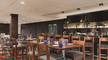 Restaurante com várias mesas e cadeiras, área de bar com prateleiras cheias de garrafas e uma parede de cor escura.