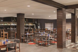 A restaurant with multiple tables and chairs, a bar area with shelves of bottles, and a modern ceiling design. - Radisson Hotel Oklahoma City Airport (Oklahoma City)