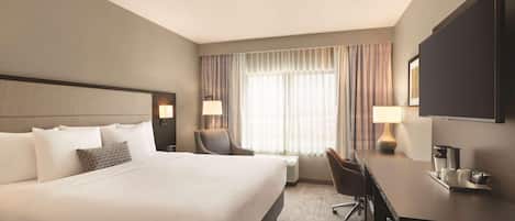 A hotel room with a large bed, a desk, two chairs, a television, and a window with curtains.