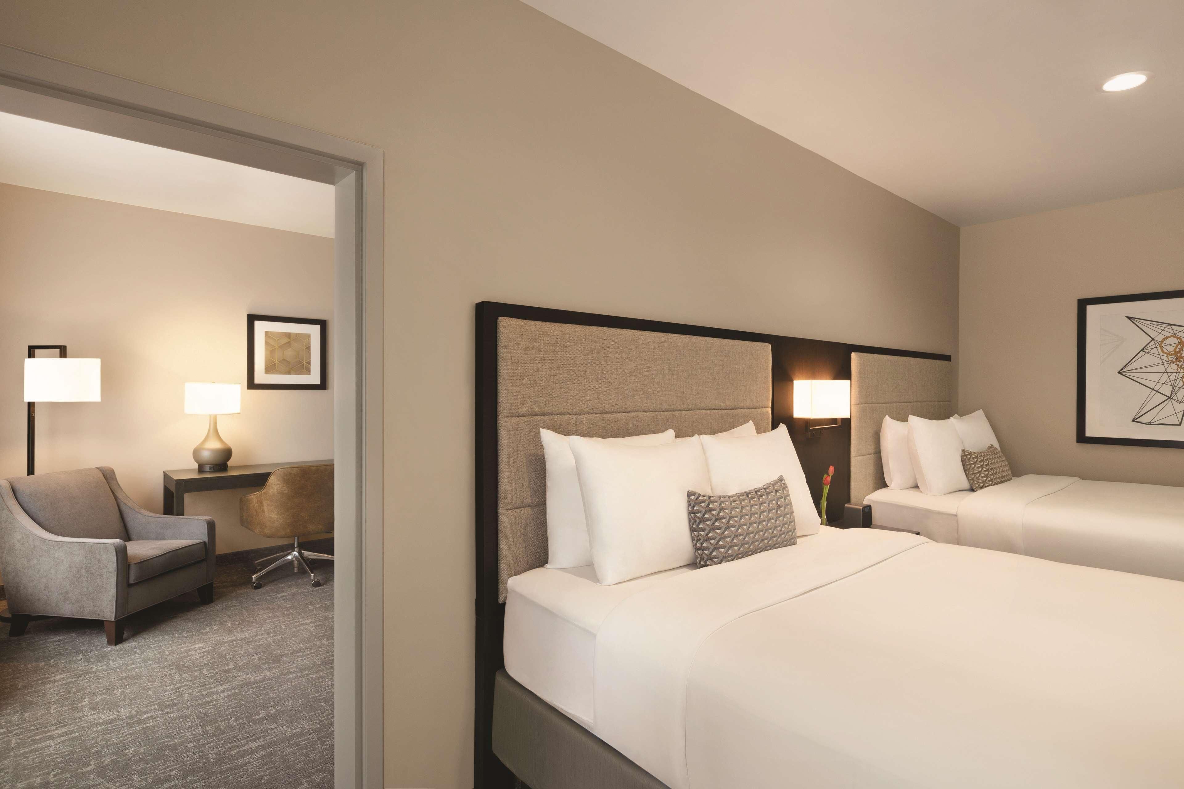 A hotel room with two beds, a desk, a chair, and a lamp.
