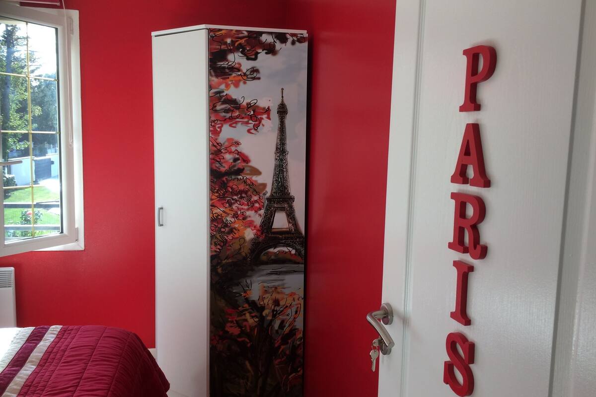 Een kamer met rode wanden, een bed met een rood-wit gestreept dekbedovertrek, een witte inbouwkast met een Parijse muurschildering, en een deur met de tekst 'PARIS' in rode letters.