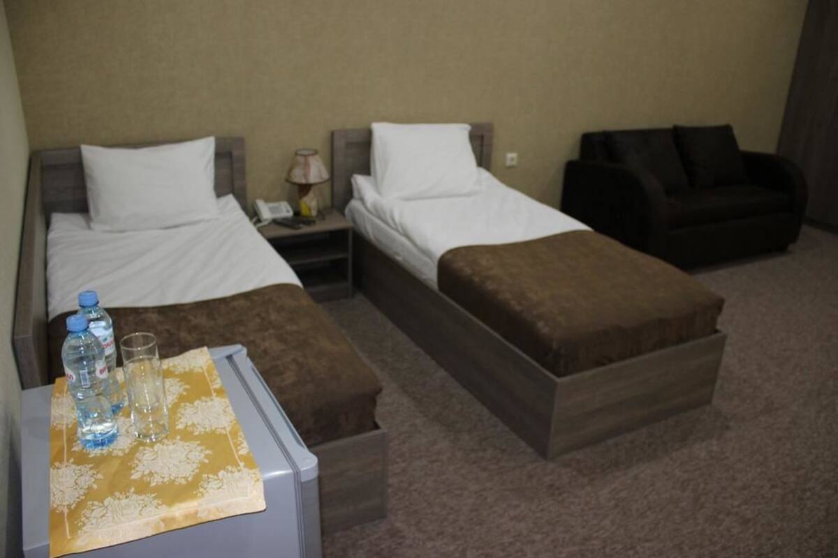 A hotel room with two single beds, a small table with a bottle and glasses, and a dark armchair.