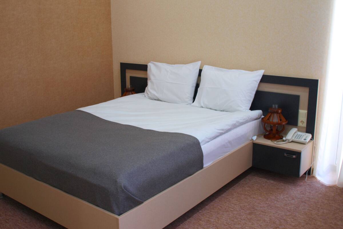 A neatly made bed with white linens and a gray throw blanket, a bedside table with a lamp and a phone, and a carpeted floor.