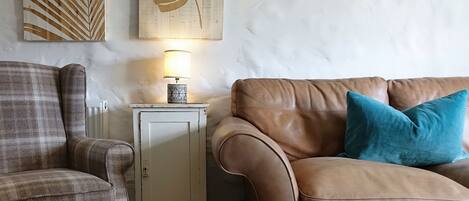 Ein Wohnzimmer mit einem braunen Ledersofa, einem karierten Sessel und einer kleinen weißen Kommode mit einer Lampe.