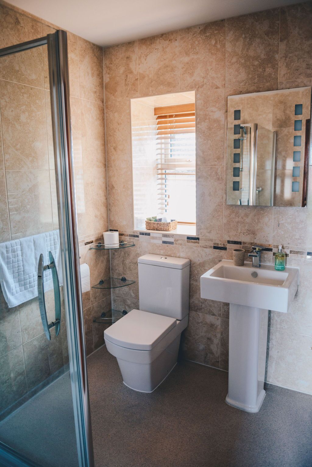 A bathroom with a glass shower enclosure, a toilet, a sink, and a window with blinds.