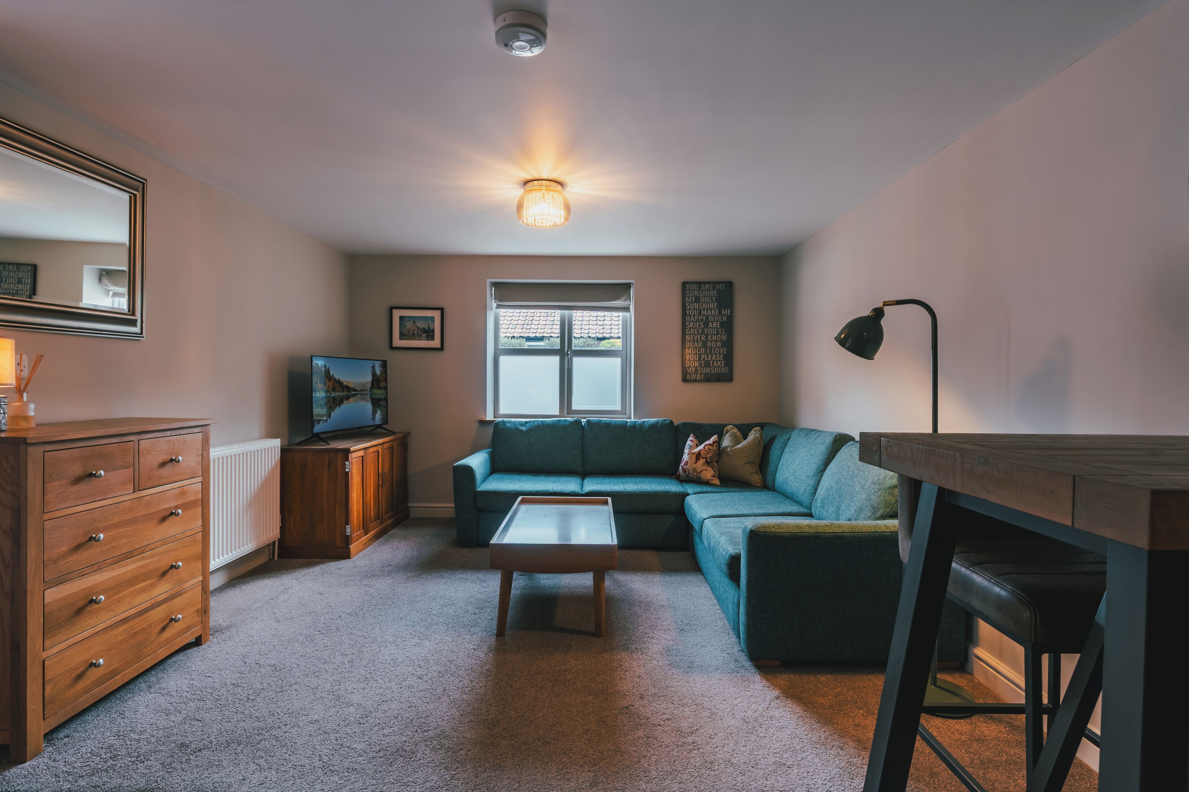 A living room with a sectional sofa, a coffee table, a wooden dresser, a TV, and a window with blinds.