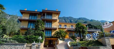 A multi-story hotel with a stone entrance, a balcony on the second floor, and a view of mountains in the background.