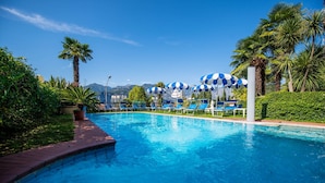 A swimming pool with blue and white umbrellas, palm trees, and a mountainous backdrop. - Hotel Cristallo (Malcesine)