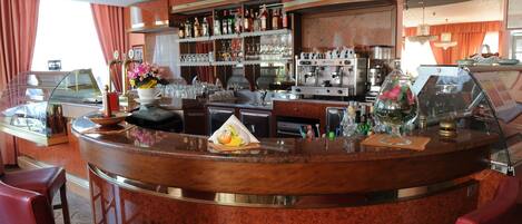 Zona de bar con mostrador de mármol, taburetes, una máquina de café y una variedad de botellas y vasos en estantes.