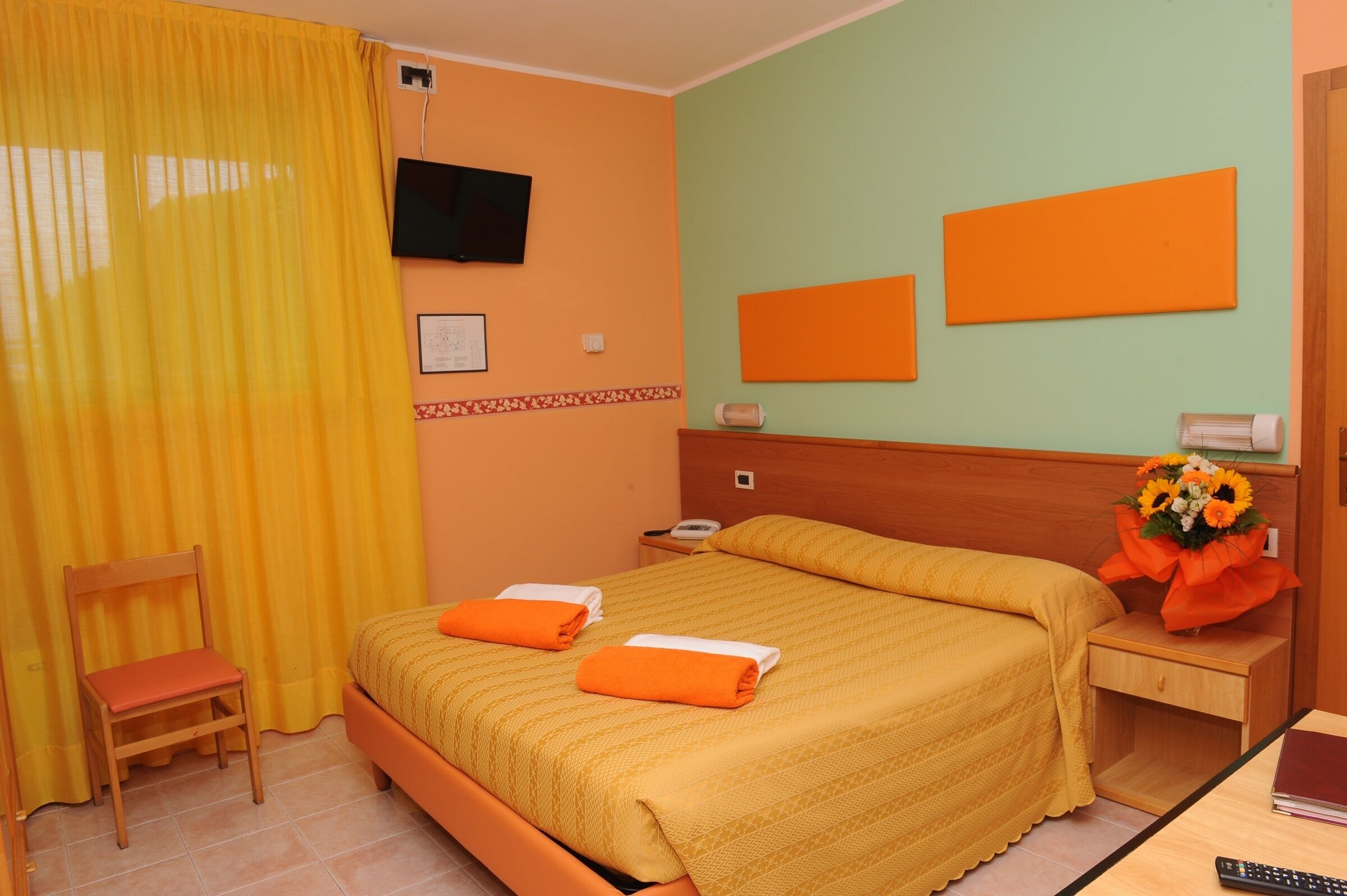 A hotel room with a bed, a chair, a bedside table with a phone, a TV on the wall, and a bouquet of flowers.