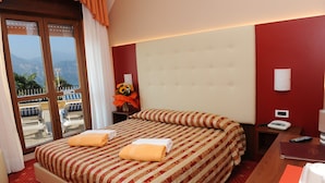 A hotel room with a large bed, a bedside table with a phone, a window with a view of mountains, and a red wall. - Hotel Cristallo (Malcesine)