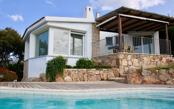 A modern house with a stone facade, large windows, and a wooden deck leading to a swimming pool.