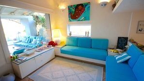 Ein Wohnzimmer mit blauem Sofa, einem Fenster mit Aussicht, einem Gemälde an der Wand und einem kleinen Tisch mit einer Lampe.