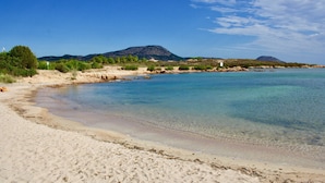 Ein Strand mit klarem Wasser, sandigem Ufer und Bergen im Hintergrund.