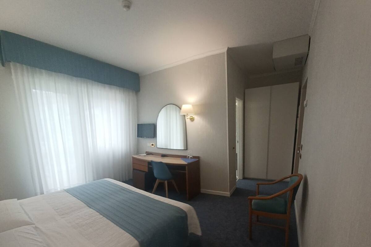 A hotel room with a bed, a desk, a chair, a mirror, and a door leading to another room.