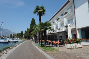 A waterfront hotel with outdoor seating, a row of palm trees, and boats in the background. - Hotel Excelsior Bay (Malcesine)