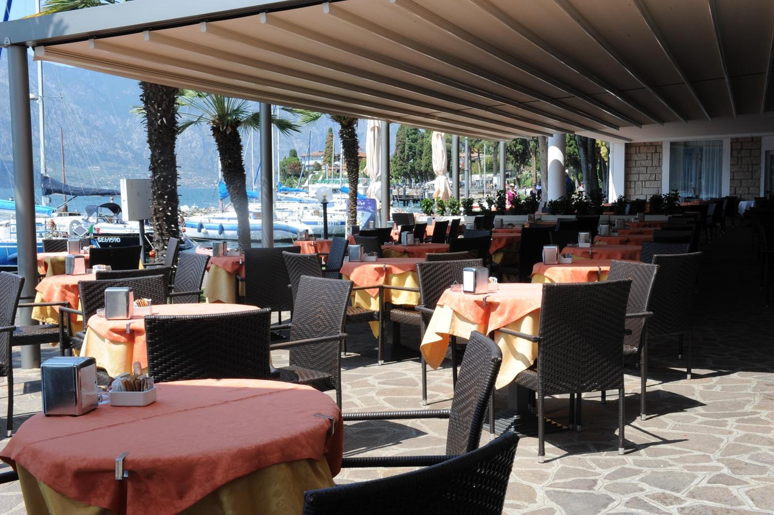 An outdoor cafe with tables and chairs, overlooking a marina with boats and mountains in the background.