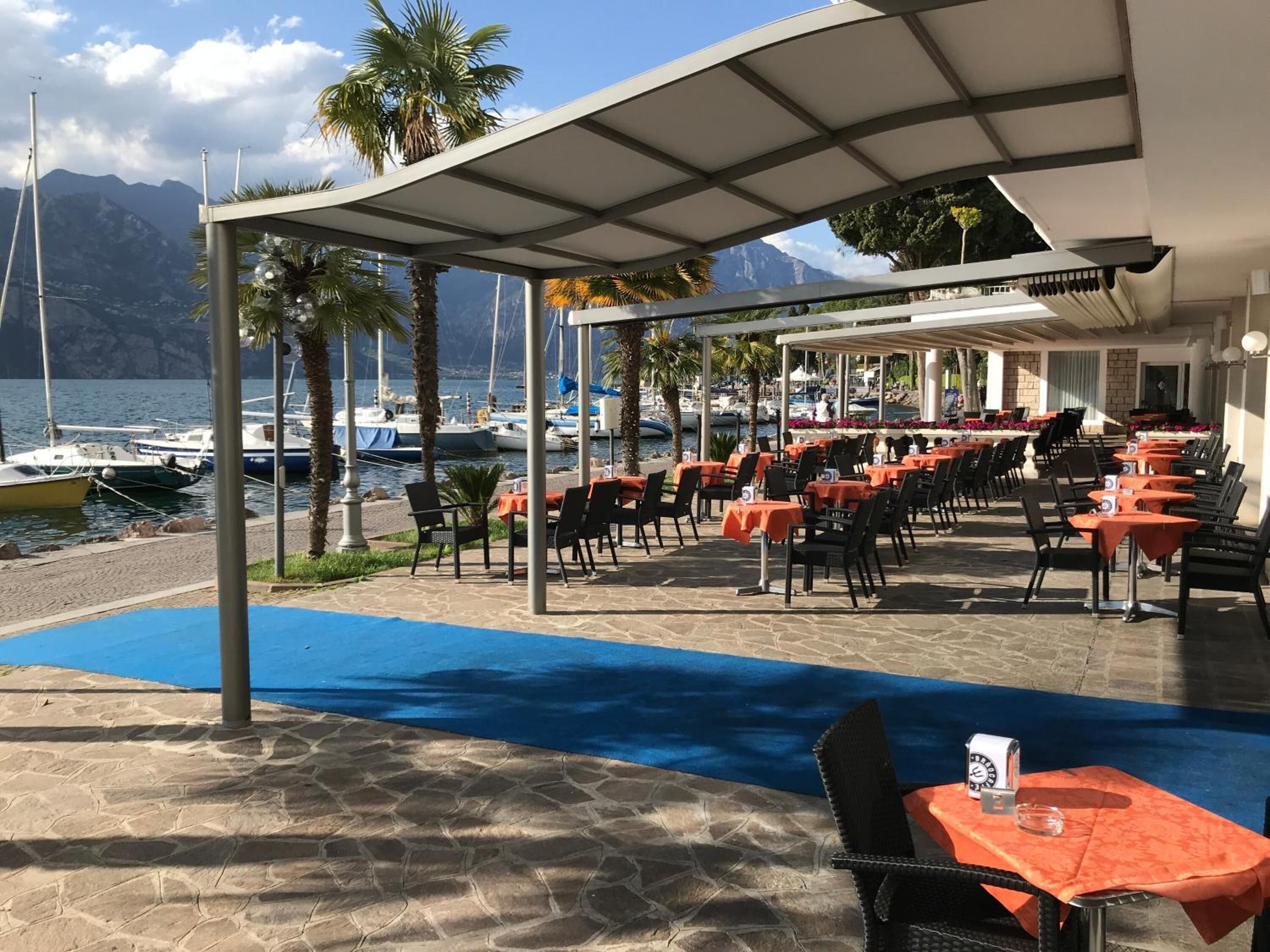 A seaside restaurant with outdoor seating, a covered patio, and a view of boats and mountains.