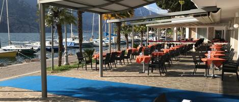 A seaside restaurant with outdoor seating, a covered patio, and a view of boats and mountains.
