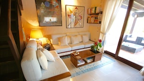 A living room with a sofa, a coffee table, and a lamp. There are bookshelves and a window with curtains. - Villa Sara (Olbia)