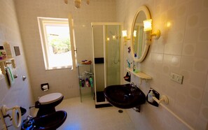 Ein Badezimmer mit Toilette, Waschbecken und Dusche. Ein Fenster sorgt für natürliches Licht und ein Spiegel ist mit Beleuchtung ausgestattet.