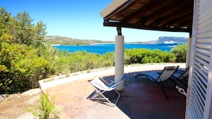A covered outdoor seating area with two lounge chairs and a table, overlooking a coastal landscape with clear blue waters and distant hills. - Villa Twin (Golfo Aranci)
