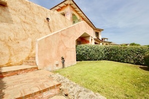 A building with a stone staircase leading to a door, a well-maintained lawn, and a hedge. - Casa Gabriella (Olbia)